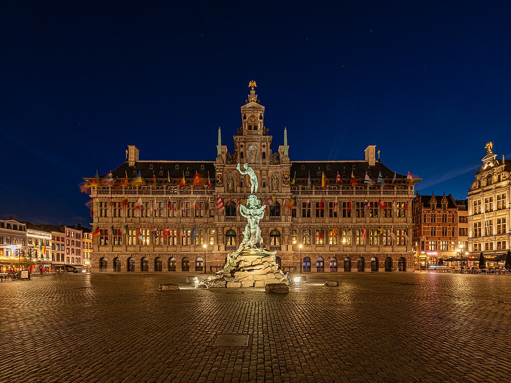 City hall of Antwerp, Belgium by night. The Brabo Fountain in the front and the hall itself have a warm yellow light against the dark blue night sky.